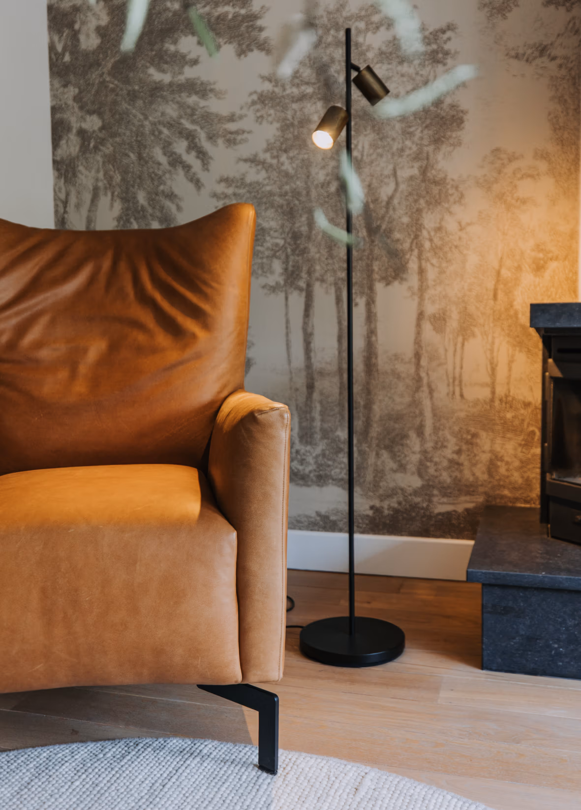Moderne woonkamerhoek met een bruine leren fauteuil, een zwarte staande lamp, een zwarte open haard en een muurschildering met een boslandschap in gedempte tinten.
