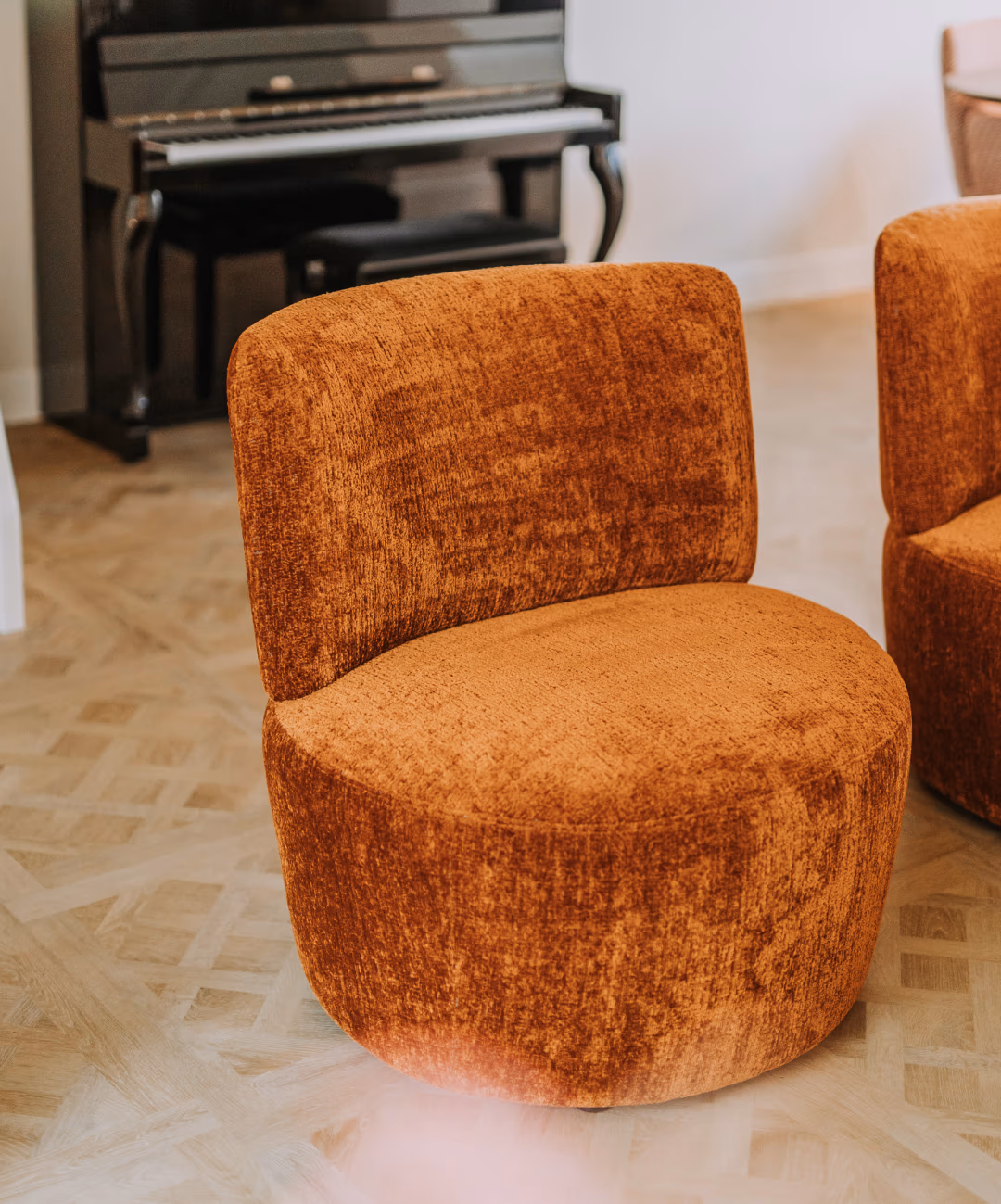 Een moderne, zachte oranje stoel met een gebogen rugleuning staat op een houten parketvloer. Een tweede, soortgelijke stoel is gedeeltelijk zichtbaar en op de achtergrond staat een zwarte staande piano.