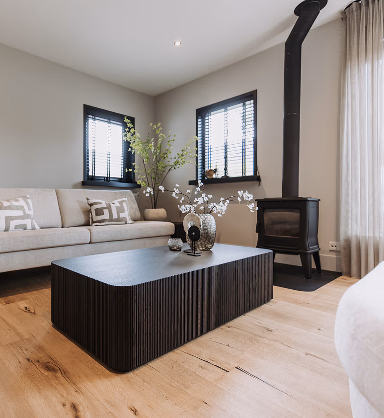 Moderne woonkamer met een beige bank, zwarte salontafel, houtkachel, houten vloer en decoraties zoals vazen en een groene plant. Twee ramen met zwarte jaloezieën en transparante gordijnen laten natuurlijk licht binnen.