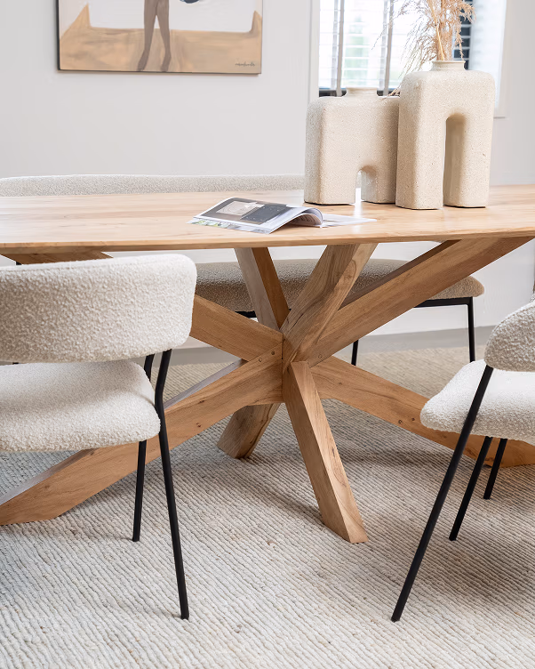 Een moderne houten eettafel met een uniek stervormig onderstel, omringd door stoelen met kussens en zwarte poten. Op de tafel staan twee beige decoratieve vazen, een tijdschrift en een klein flesje. De kamer heeft een neutrale inrichting.