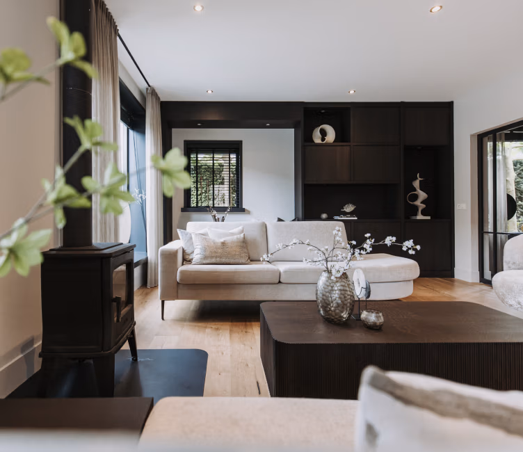 Een moderne woonkamer met een beige bank, donkere houten planken, een zwarte houtkachel en grote ramen. Op de voorgrond staat een houten salontafel met decoratieve vazen en takken.